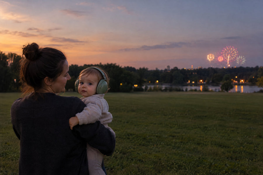Parent holding a baby wearing soft padded earmuffs at the far edge of a park at dusk, with fireworks far away over the trees