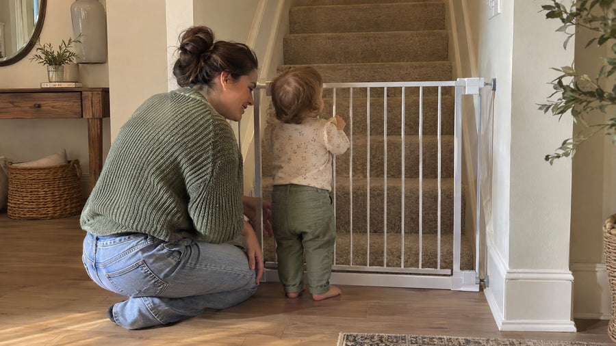Toddler holding a baby gate fitted at the bottom of the stairs, with a parent kneeling beside
